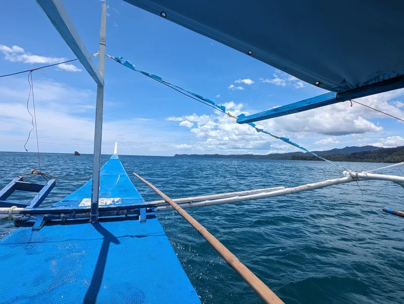 First-person view from the side of a blue bangka outrigger boat looking out over clear blue ocean toward a lush green coastline under a bright sky near Sabang, Palawan, Philippines