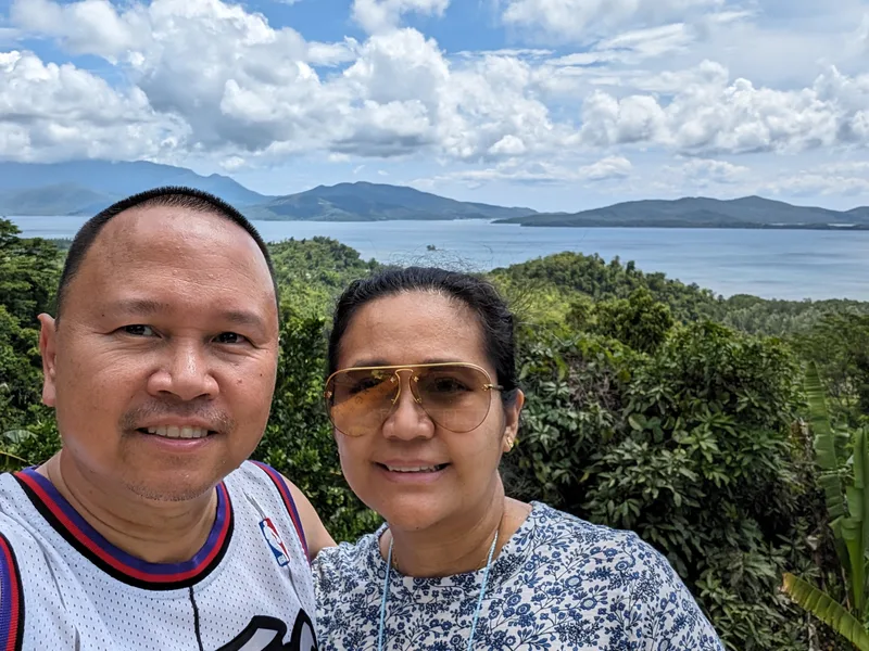 Couple selfie with a panoramic view of a bay with islands and lush green hills in the background under a bright blue sky with white clouds Palawan Philippines