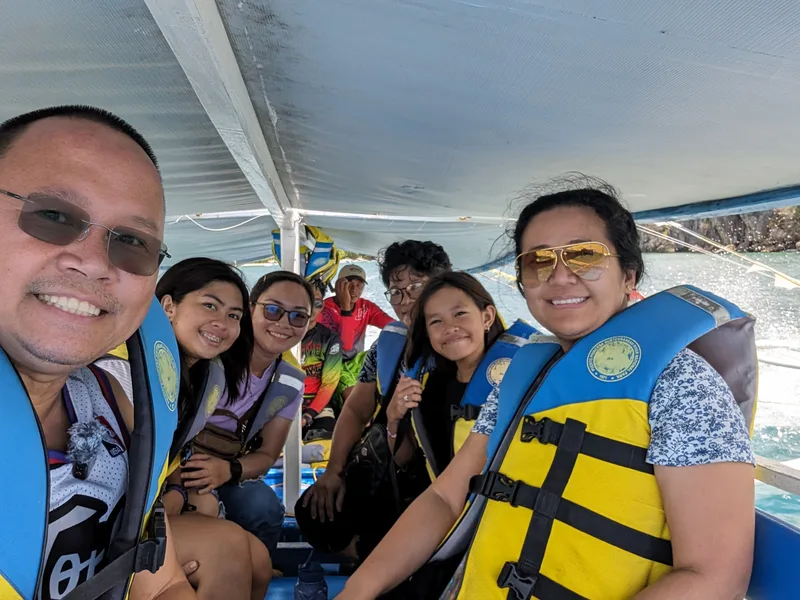 Group selfie inside a bangka boat with a man and woman in the foreground wearing life vests smiling at the camera and other family members behind them with ocean and land in the background Sabang, Palawan, Philippines