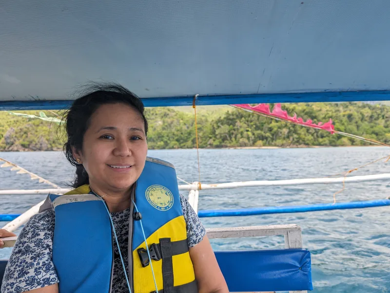 A woman in a blue and yellow life vest smiling on a bangka boat with lush green hills and calm ocean in the background near Sabang, Palawan, Philippines