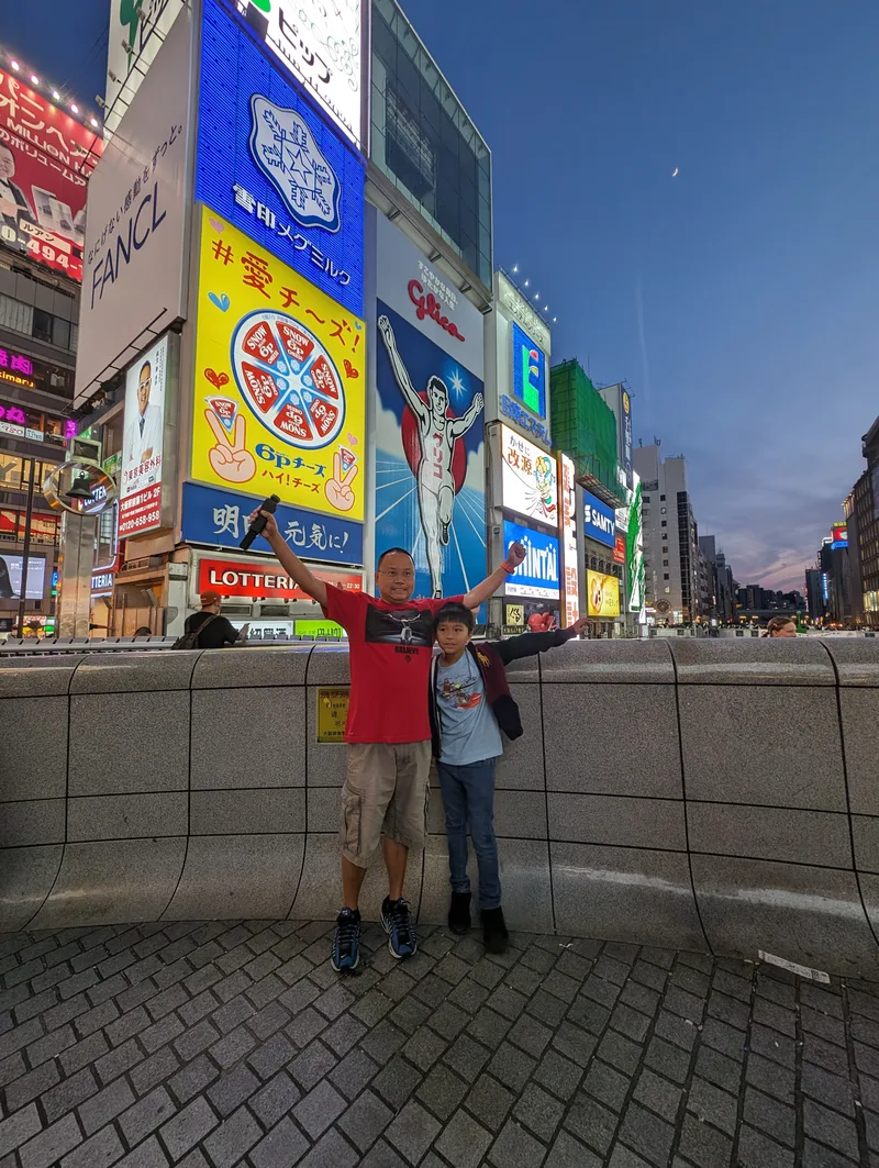 Father and son with arms raised mimicking the iconic Glico Man pose in front of the Glico Man sign in Dotonbori, Osaka, Japan.