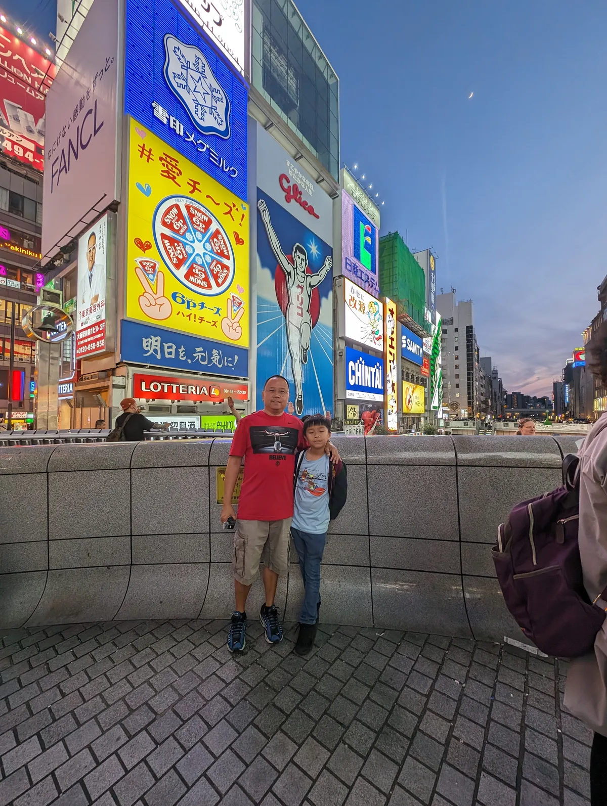 Father and son posing in front of the iconic Glico Man sign with neon billboards in Dotonbori, Osaka, Japan.