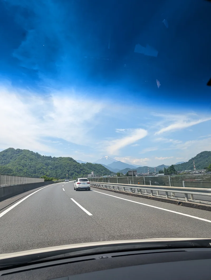 Mount Fuji visible from a highway in Yamanashi, Japan, flanked by green mountains and blue sky.