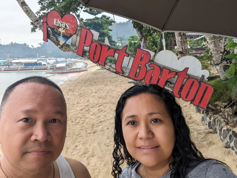Couple selfie in front of the I Love Port Barton sign on the beach with traditional outrigger boats anchored in the bay behind them