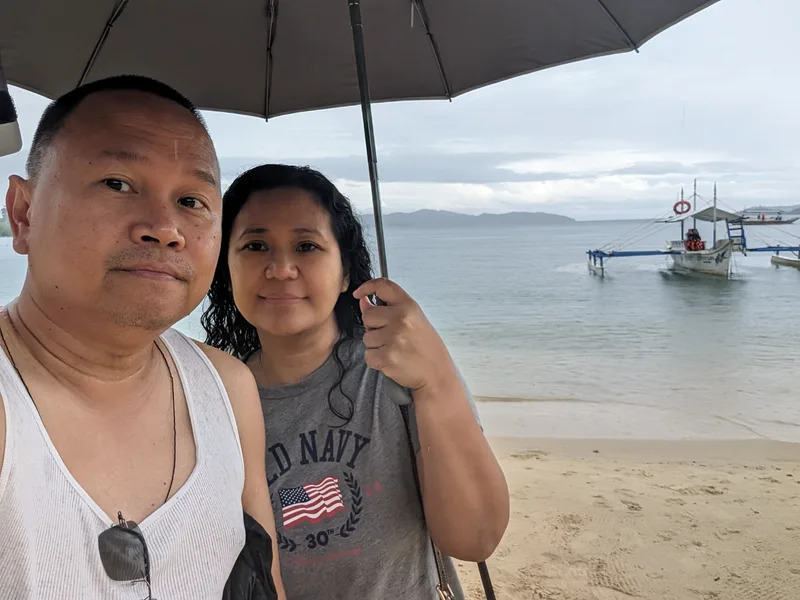 Couple selfie on Port Barton beach in Palawan Philippines with traditional outrigger boats anchored in the background on calm grey water