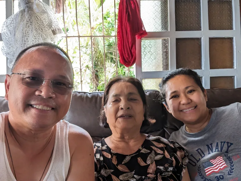 Selfie of man, woman, and elderly woman smiling on a couch indoors with a window and green foliage in the background at a family home in Caramay Roxas Palawan Philippines
