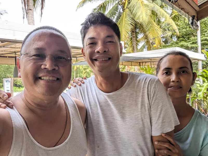 Three people smiling for a selfie in a tropical outdoor setting with palm trees and greenery in Palawan Philippines