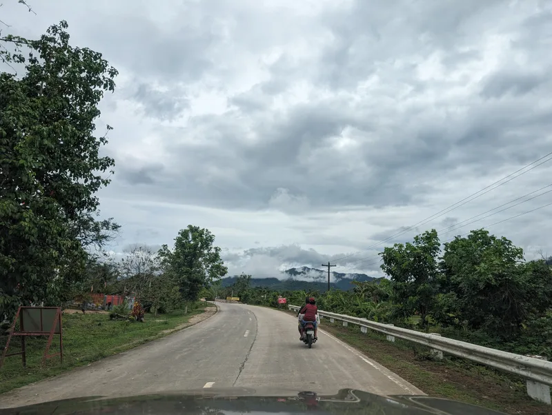 Winding rural road in Palawan Philippines viewed from a car with a motorcycle ahead and lush green trees and misty mountains in the background