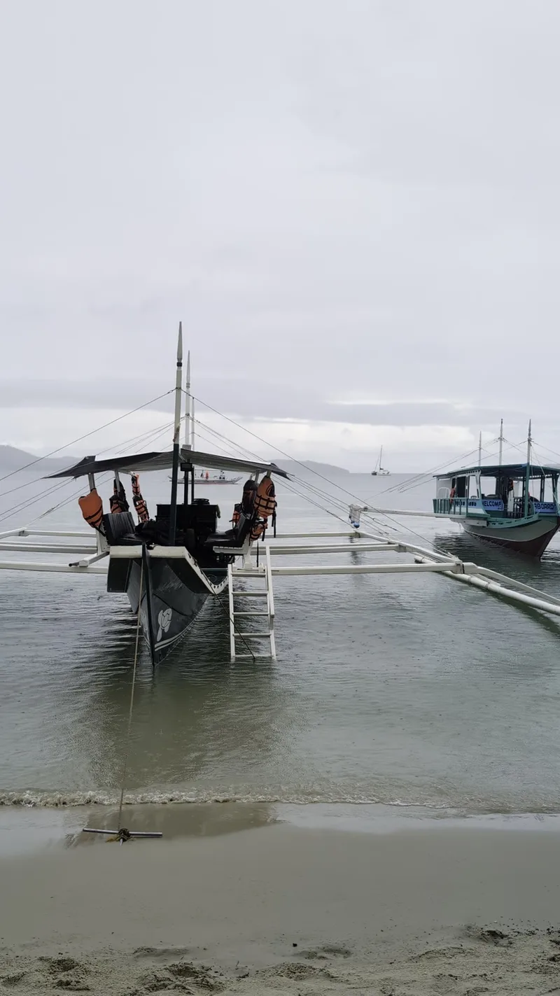 Two traditional Filipino outrigger boats anchored in shallow calm water near Port Barton beach Palawan under an overcast sky